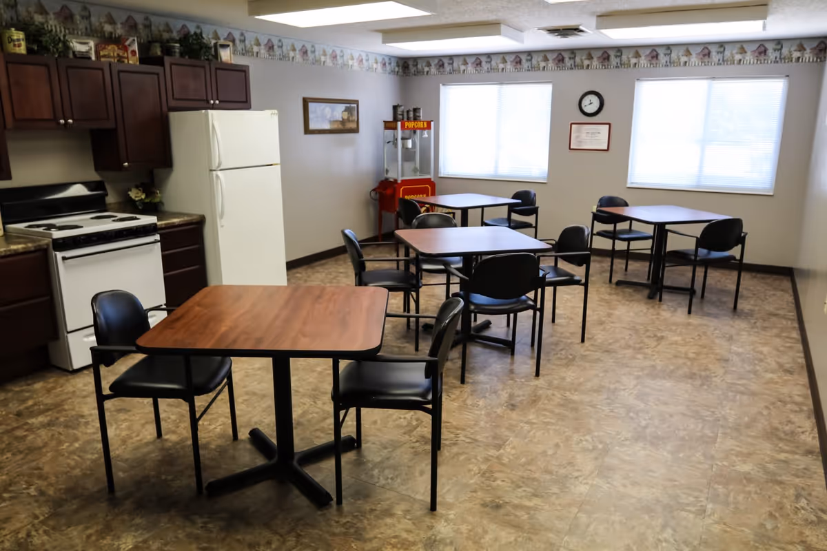 Communal dining area with several tables and chairs and a small kitchen with a stove and refrigerator.
