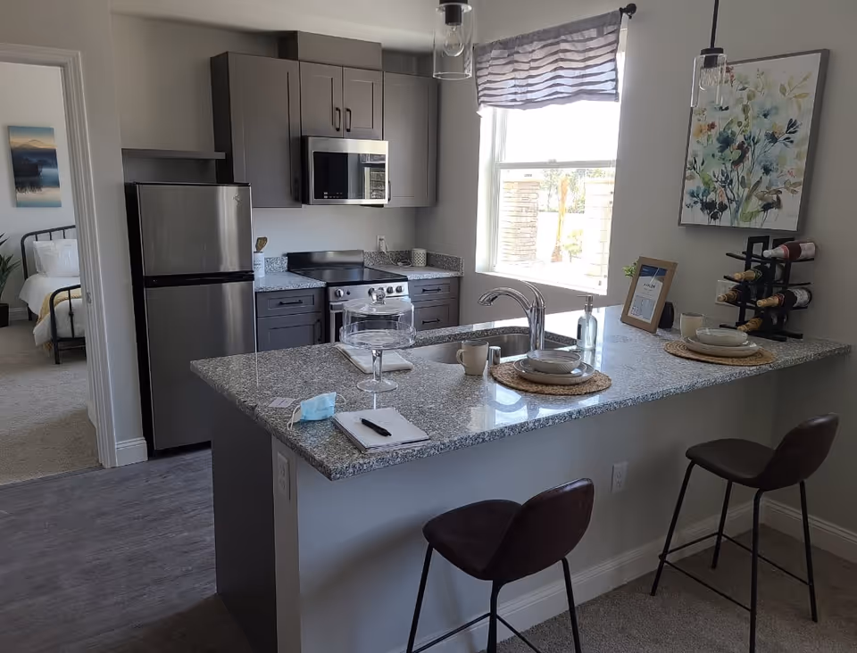 Modern kitchen area with gray cabinets, stainless steel refrigerator, microwave, and stove. A granite countertop island with two brown bar stools, place settings, a cake stand, and a face mask on it. A window with a striped curtain lets in natural light. A bedroom with a bed and artwork is visible through an open doorway.