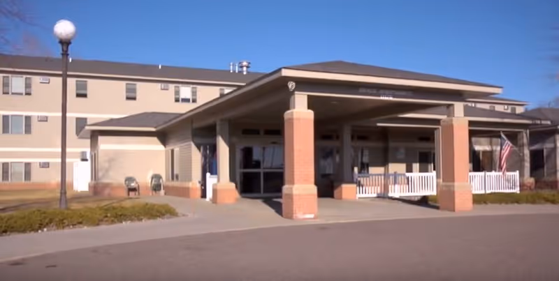 Exterior view of Shepherd of Grace Senior Community building entrance with a covered drop-off area supported by brick columns, a white picket fence, an American flag, and a street lamp on the left side under a clear blue sky.