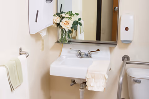 A clean bathroom sink area with a white sink, a mirror above it, a vase with white and peach flowers on the sink, a towel hanging on the sink, a towel rack with a green towel, a soap dispenser mounted on the wall, and a toilet partially visible to the right with a grab bar beside it.