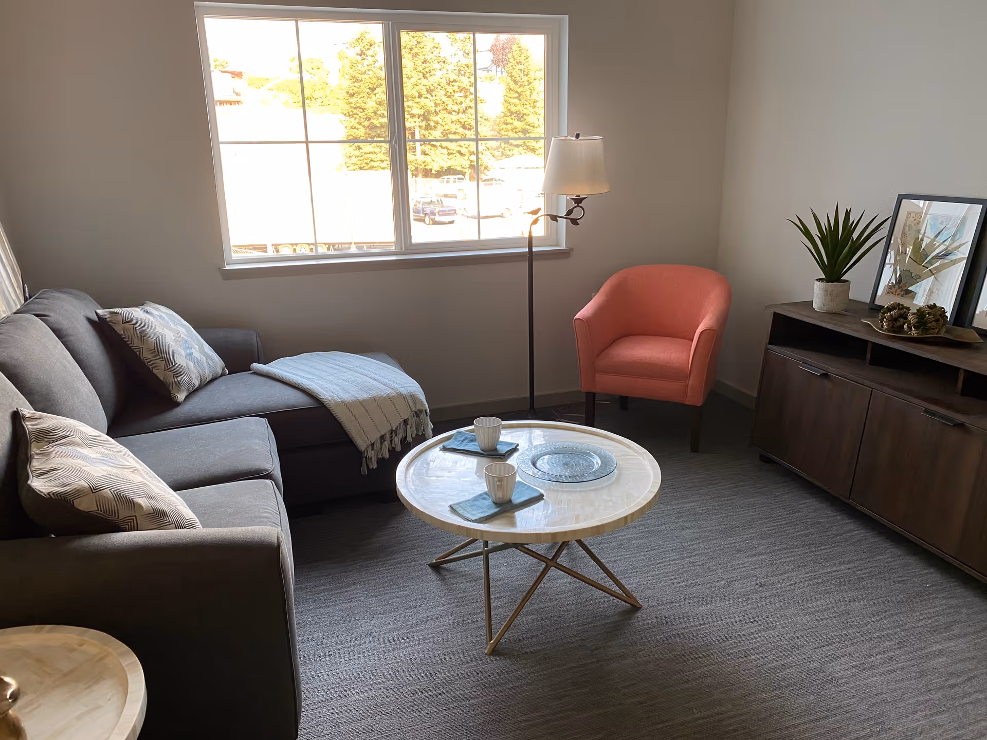 A cozy living room with a gray sectional sofa adorned with patterned pillows and a light blue throw blanket. In front of the sofa is a round coffee table set with two cups on napkins and a decorative plate. To the right, there is a coral-colored armchair next to a floor lamp. A dark wooden cabinet with potted plants and framed artwork is against the wall. A large window lets in natural light, showing trees and parked cars outside.