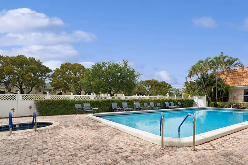 Outdoor swimming pool with clear blue water surrounded by a brick-paved deck. Several lounge chairs are lined up along a white fence with green bushes and trees behind it. A small circular hot tub is visible on the left side. The sky is partly cloudy with blue patches.