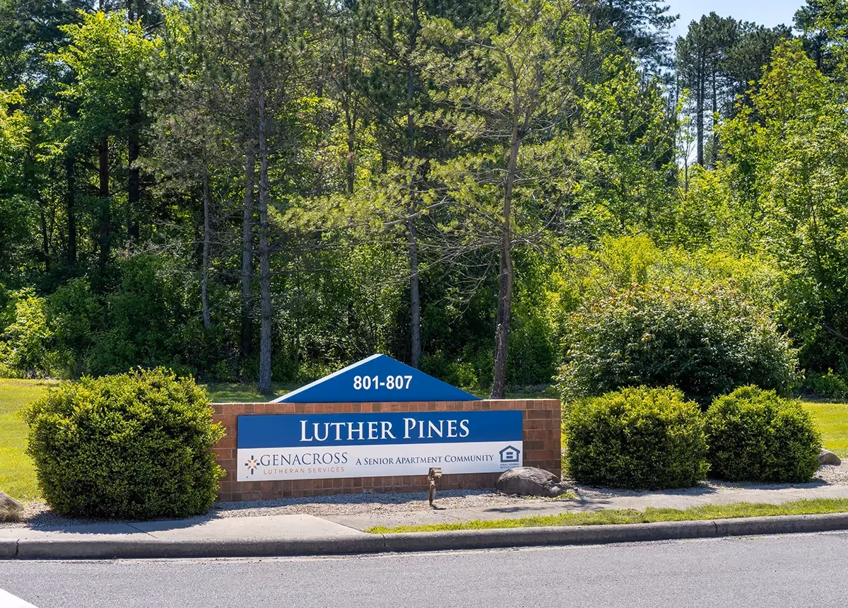 Outdoor view of a brick and blue sign for Luther Pines, a senior apartment community by Genacross Lutheran Services, surrounded by green bushes and trees.