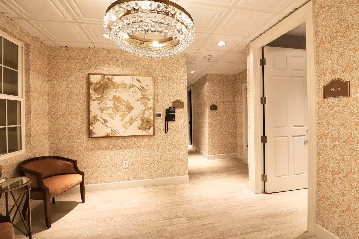 Interior hallway of a senior living facility with patterned wallpaper, a crystal chandelier, a framed abstract artwork on the wall, a brown upholstered chair next to a small glass-top table, and a door labeled 'Nails'.