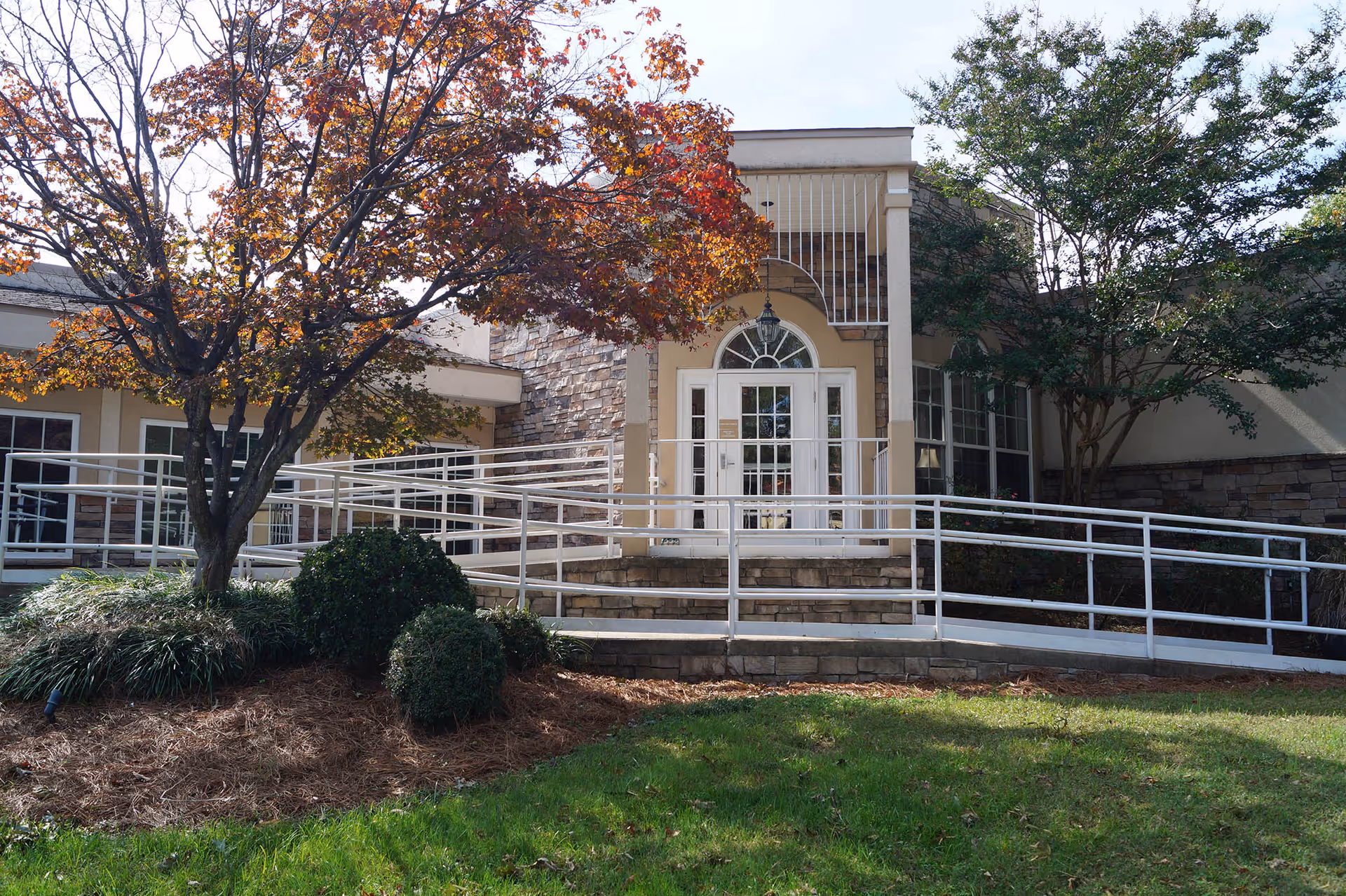 Exterior view of a building entrance with a white door and arched window above it, surrounded by stone and beige walls. There is a white metal ramp with railings leading up to the entrance. Trees with autumn-colored leaves and green bushes are in the landscaped area in front of the building.