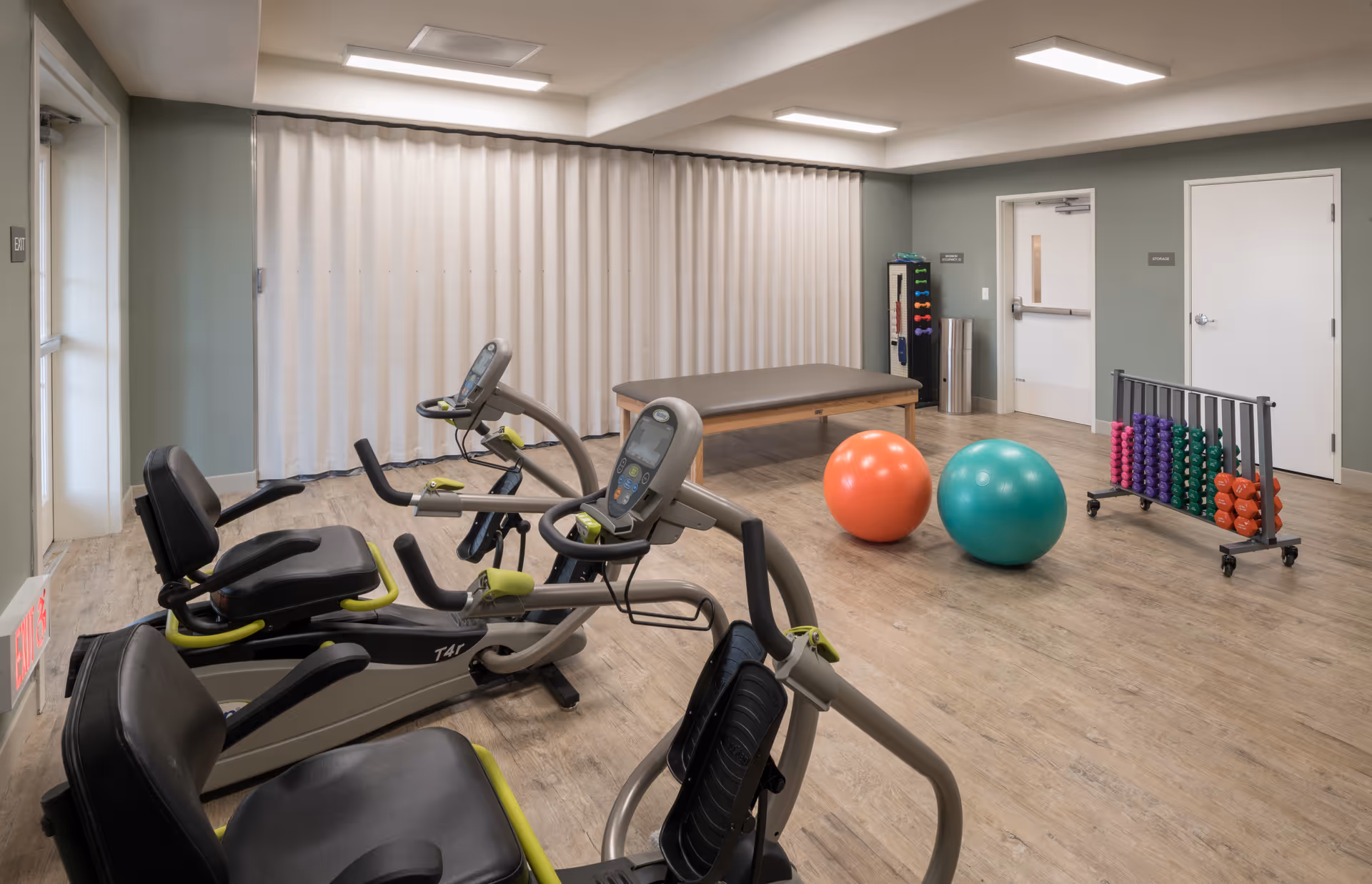 A fitness room with two recumbent exercise bikes, a treatment table, two large exercise balls (one orange and one teal), and a rack of colorful dumbbells against a light green wall with two white doors.