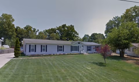 Single-story senior care building with light siding, a manicured lawn, and trees.