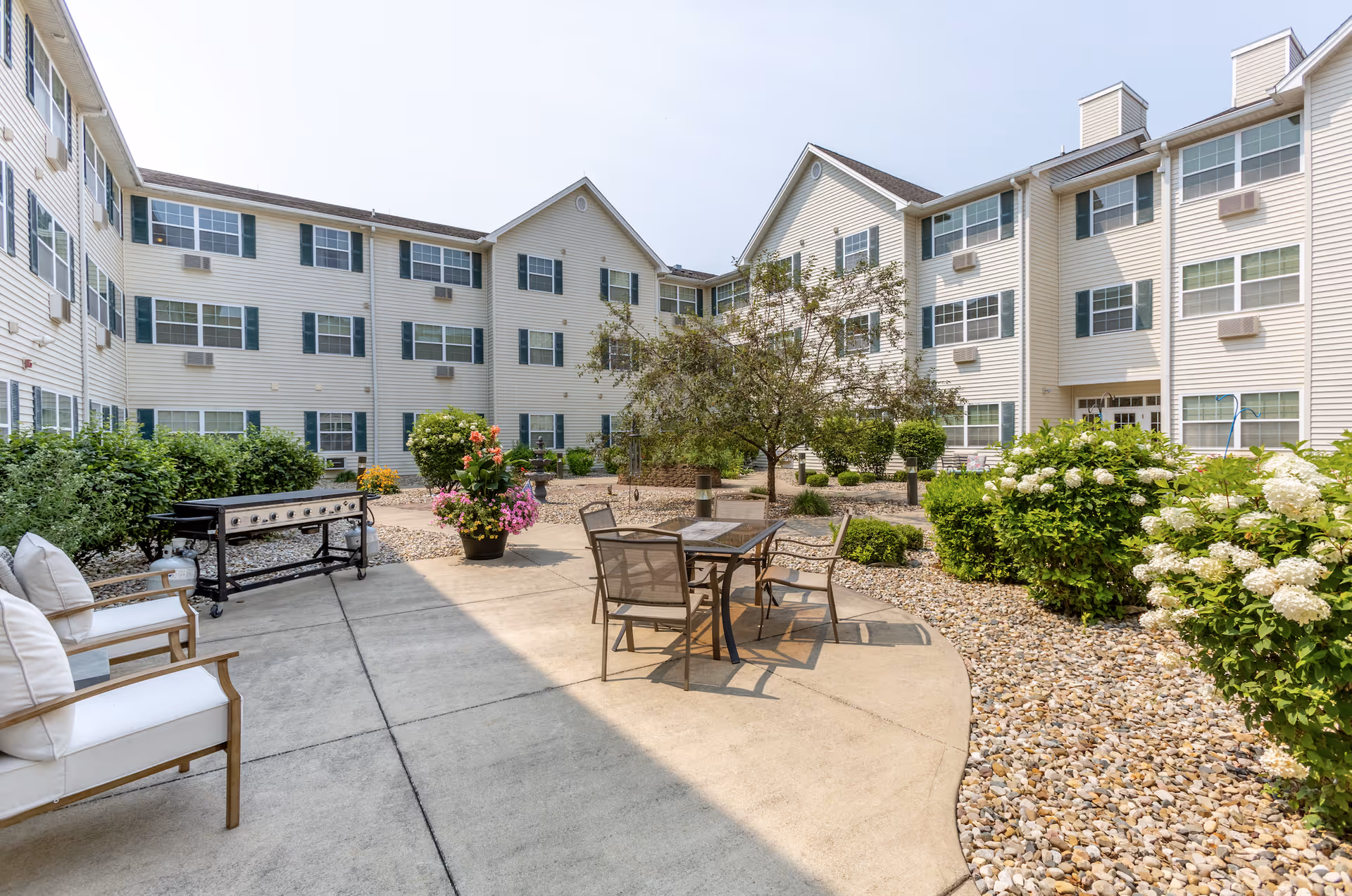 Outdoor courtyard area of a senior living facility with a concrete patio, outdoor seating including cushioned chairs and a table with chairs, a large grill, and landscaped garden beds with bushes, flowers, and a tree. The building surrounding the courtyard is three stories tall with many windows and beige siding.