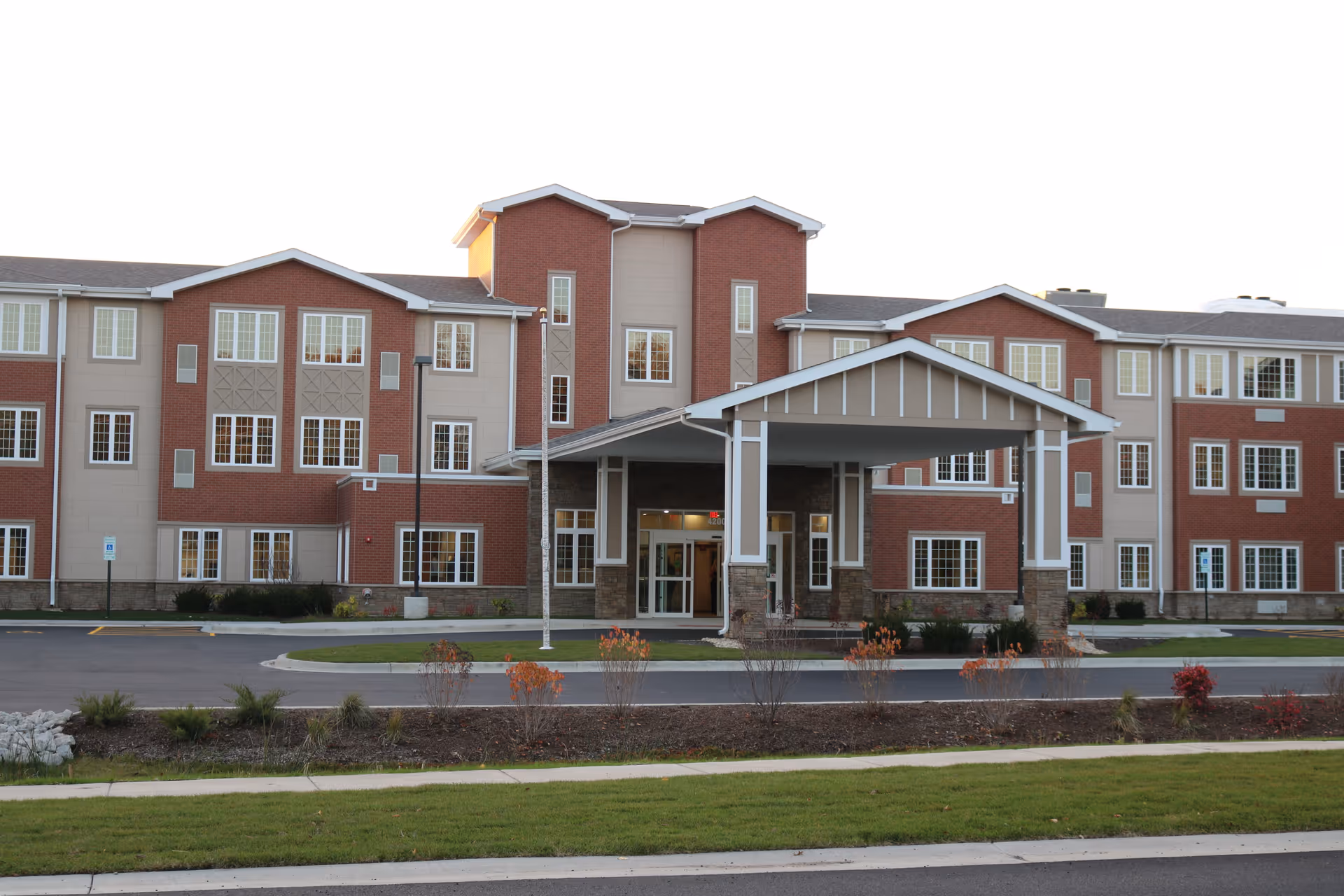 Front exterior view of a three-story supportive living facility building with a covered entrance, multiple windows, and landscaped grounds with grass and small shrubs.