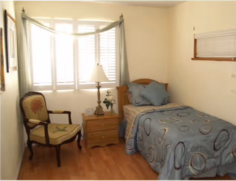 Small bedroom with a twin bed, nightstand and lamp, an upholstered chair, and a window with shutters.