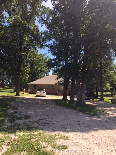 Single-story tan building with a front porch, two parked cars, and large trees lining a gravel driveway.