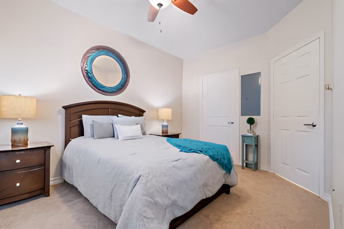 A bedroom with a large bed covered in light gray bedding and a teal throw blanket. There are two matching wooden nightstands with lamps on either side of the bed. A round decorative mirror with a blue frame hangs on the wall above the headboard. The room has beige carpet, white walls, a ceiling fan, and two white doors on the right side.