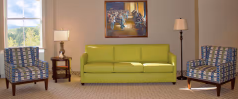 A living room area with a green three-seat sofa centered between two patterned armchairs. There is a small wooden side table with a lamp on the left side of the sofa and a tall floor lamp on the right. A large window on the left side lets in natural light, and a framed painting hangs on the wall above the sofa.