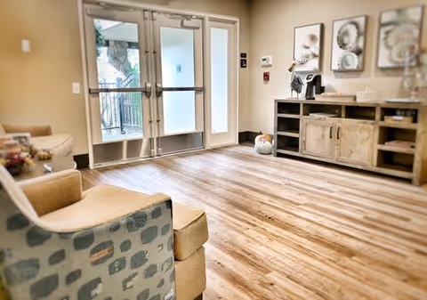 Sunny lobby/common seating area with armchairs, a wooden console and artwork facing double glass entry doors.