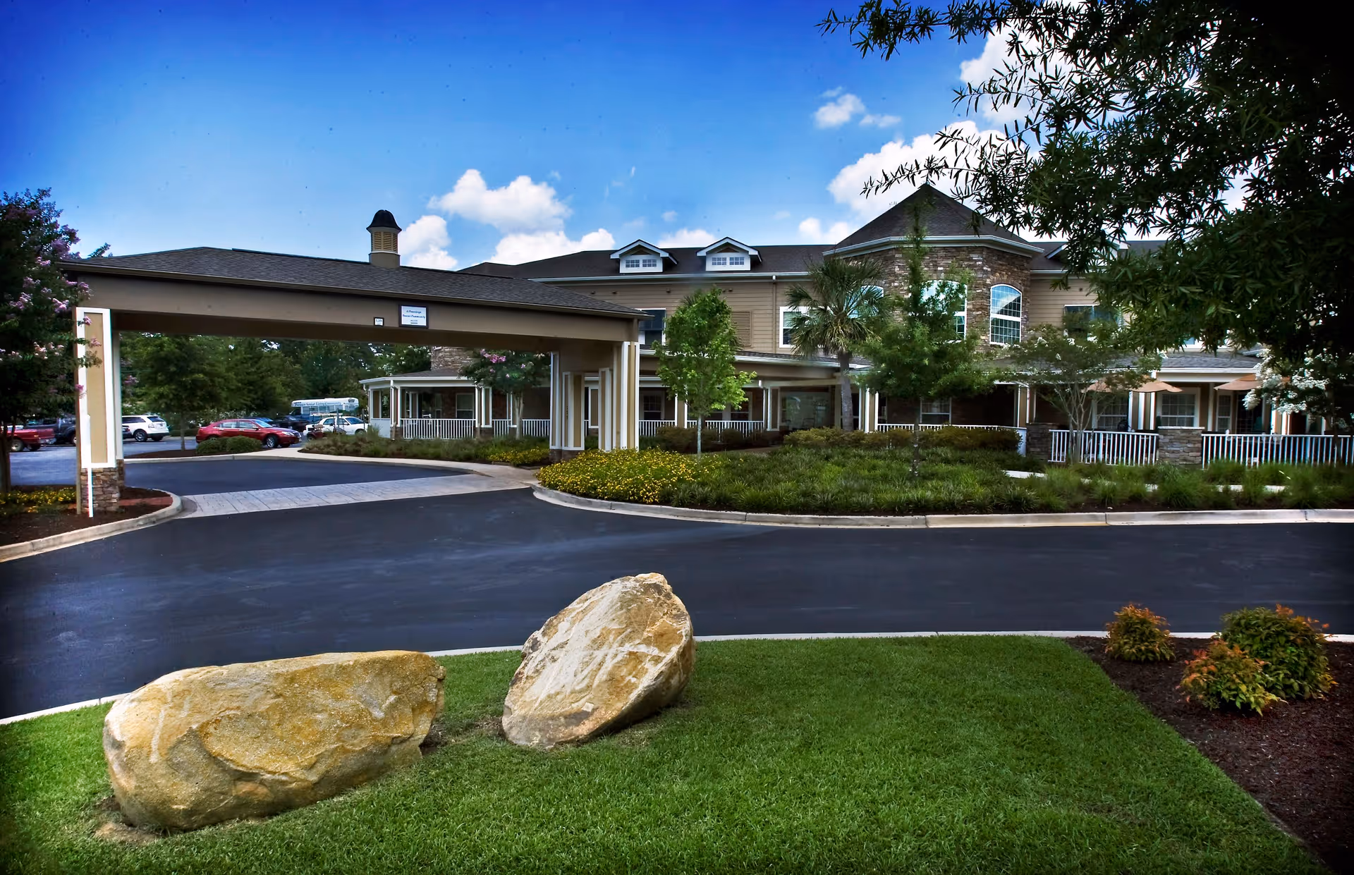 Exterior view of The Village at Southlake senior living facility showing a driveway with a covered entrance, landscaped greenery, large rocks on a grass lawn, and a multi-story building with windows and a porch in the background under a blue sky with some clouds.