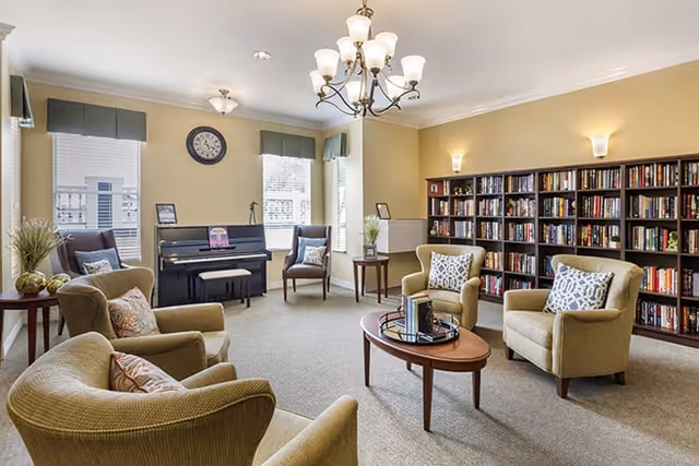 Bright communal living room with upholstered armchairs arranged around a coffee table, bookshelves along the wall, and a piano beneath windows.