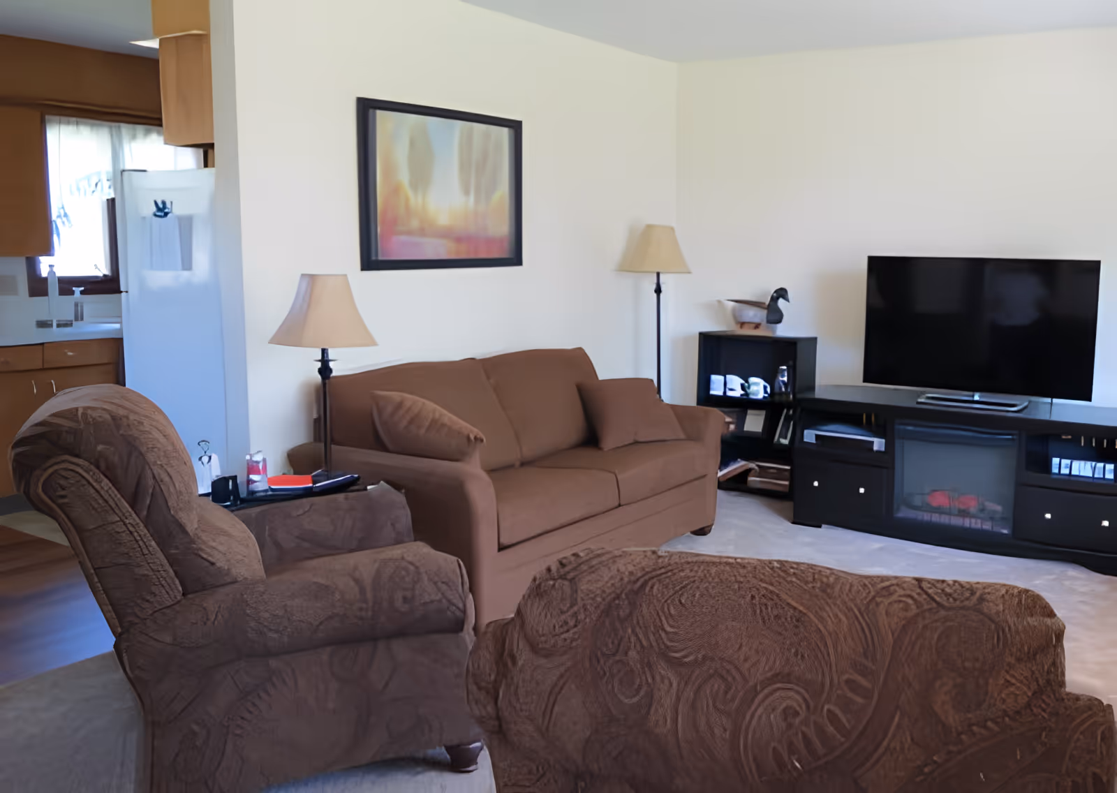 A cozy living room with a brown sofa and two patterned armchairs. There are two lamps, one floor lamp and one table lamp, on either side of the sofa. A framed landscape painting hangs on the wall above the sofa. A black entertainment center with a flat-screen TV and decorative items is positioned against the wall. The kitchen area with wooden cabinets and a white refrigerator is partially visible in the background.