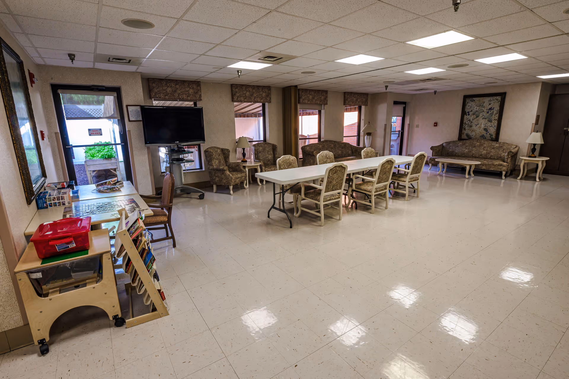 A spacious common room in a senior living facility with a long white table surrounded by cushioned chairs in the center. There are several upholstered armchairs and sofas along the walls, a flat-screen TV mounted on a stand near the windows, and a small table with books and games on the left side. The room has a tiled floor, fluorescent ceiling lights, and windows with valances letting in natural light.