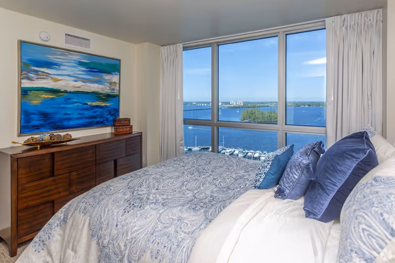 A bedroom with a large window offering a view of a marina and water. The bed is covered with a blue and white patterned comforter and several blue pillows. A wooden dresser with decorative items and a large abstract painting in blue and green tones is positioned against the wall.