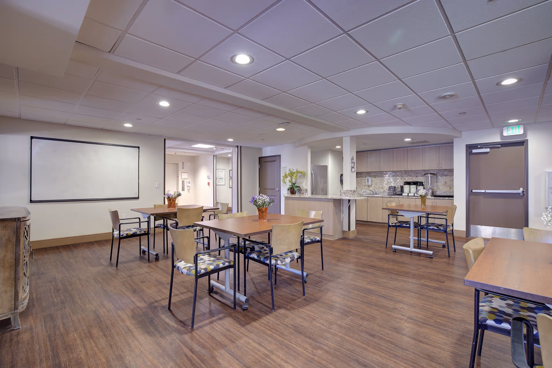 A spacious, well-lit common area with several tables and chairs arranged for group seating. The room features wood flooring, a whiteboard on one wall, and a kitchenette area with cabinets, a countertop, and appliances in the background. Small flower arrangements are placed on the tables, and recessed ceiling lights illuminate the space.