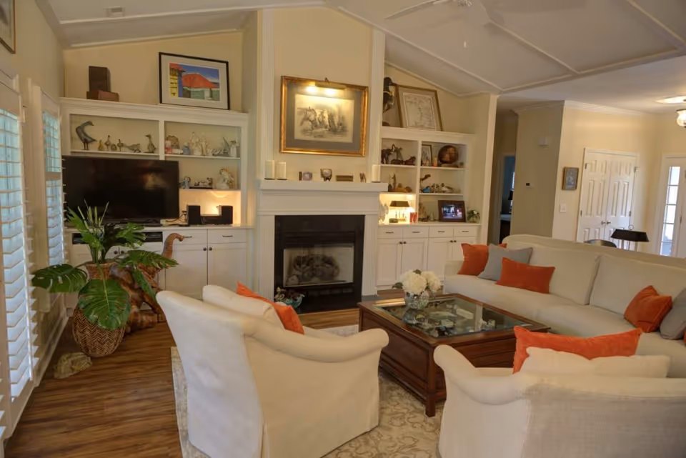 A cozy living room with a fireplace flanked by built-in shelves, a TV, a large sectional sofa and armchairs around a wooden coffee table, and decorative plants and artwork.