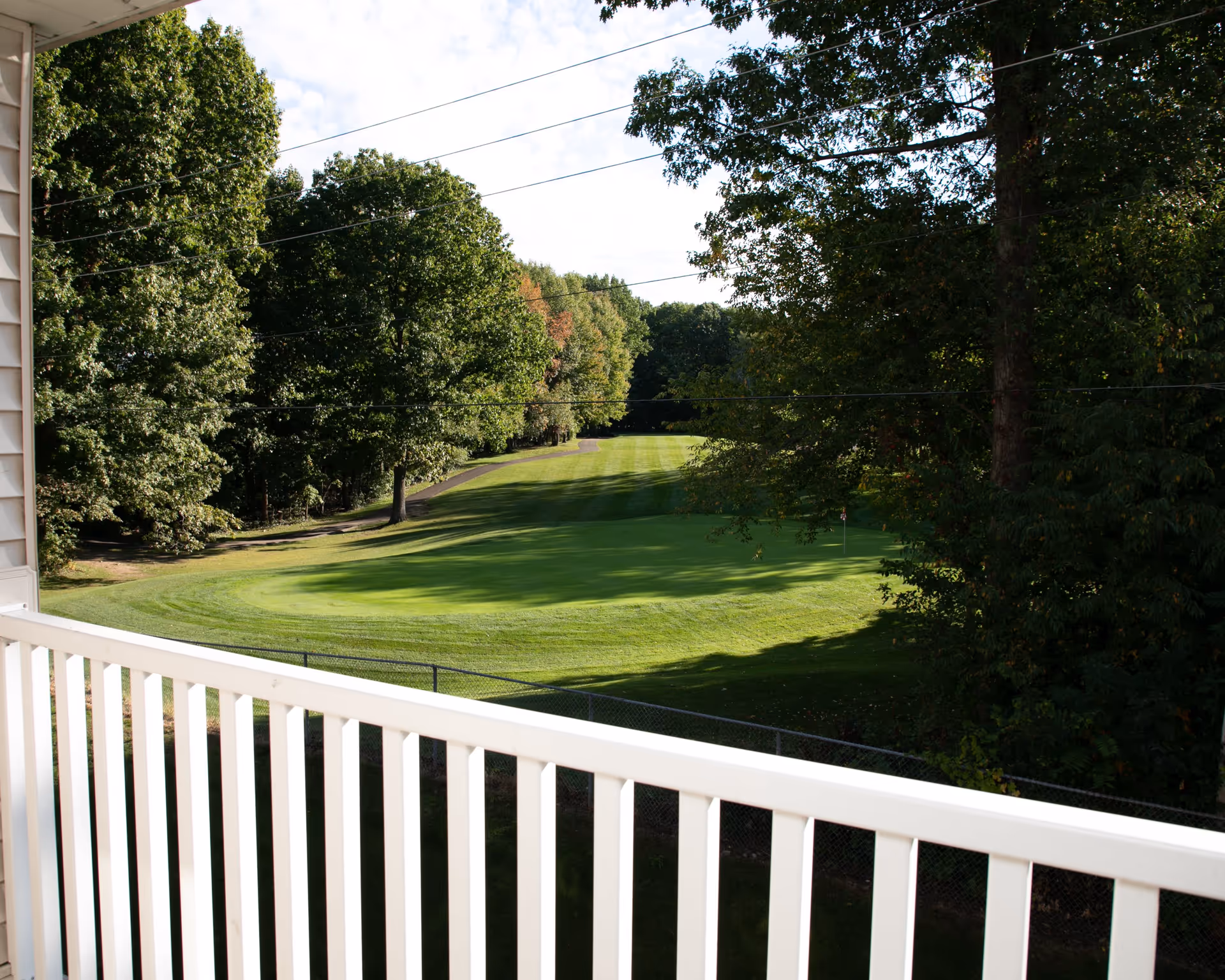 View from a balcony with white railing overlooking a well-maintained green lawn area surrounded by trees under a partly cloudy sky.
