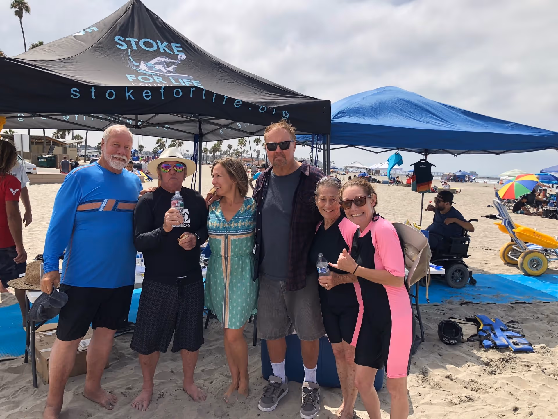 A group of six adults standing together on a sandy beach under two canopy tents. Some are wearing wetsuits and holding water bottles, while others are dressed casually. The background shows more people, beach umbrellas, and palm trees under a cloudy sky.