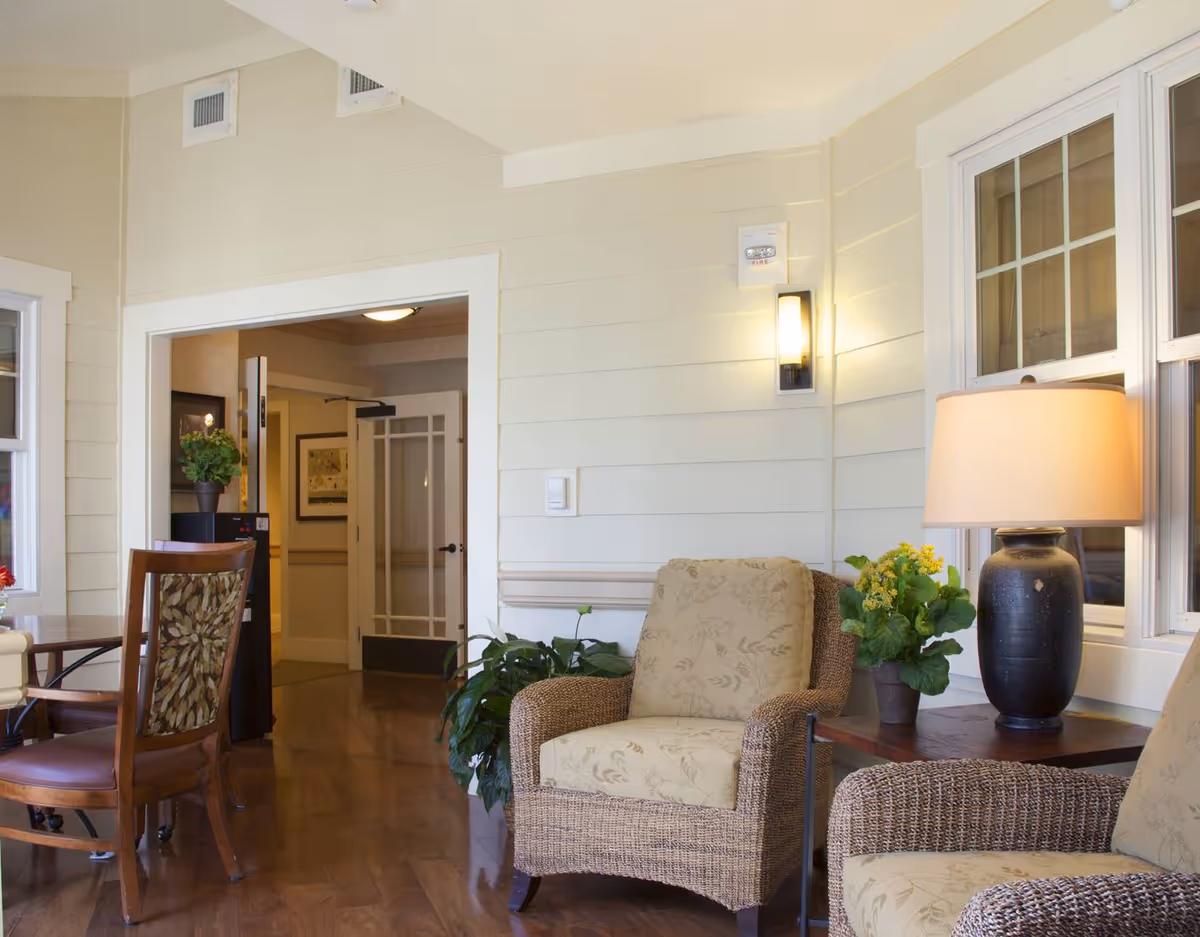 A cozy interior seating area with wicker armchairs featuring floral cushions, a wooden side table with a black lamp and a potted plant, and a wooden chair with a floral backrest near a table. The room has light-colored walls, wooden flooring, and a doorway leading to another room with a glass-paneled door.