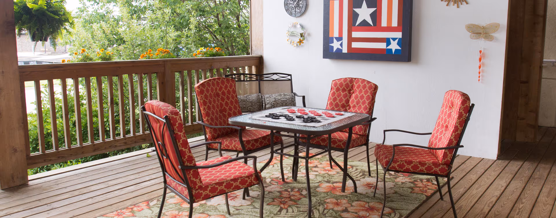Covered outdoor patio area with a wooden floor and railing, featuring a square table with a checkerboard game set up on it, surrounded by four red cushioned chairs. There is a floral patterned rug under the table and chairs. The white wall behind has decorative items including a framed artwork with stars and stripes, a clock, and a dragonfly ornament. Greenery and flowers are visible outside the railing.