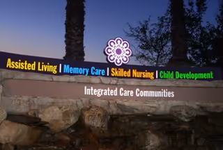 Illuminated sign at dusk for Integrated Care Communities with categories Assisted Living, Memory Care, Skilled Nursing, and Child Development displayed in different colors, set against a backdrop of palm trees and a darkening sky.