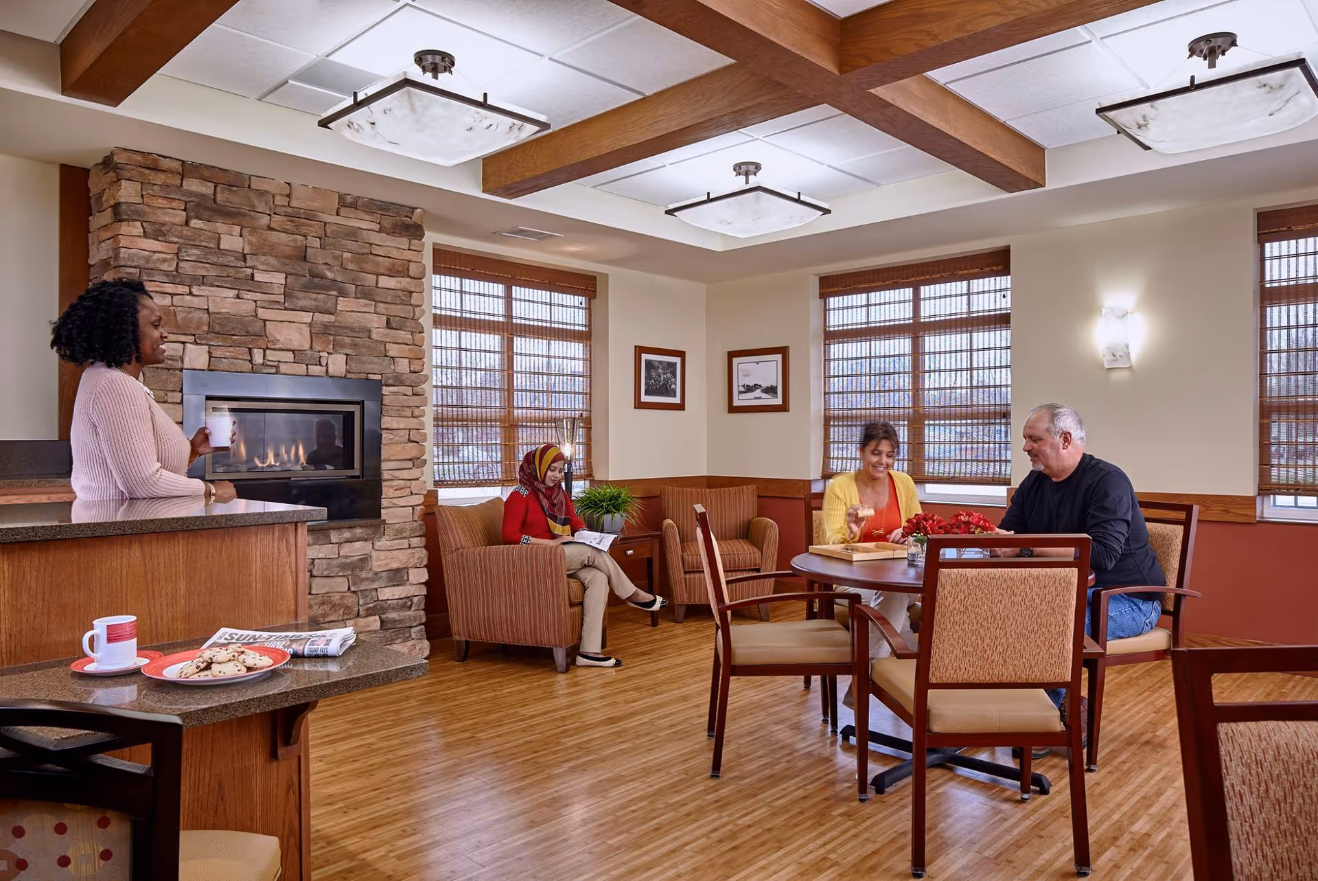 A cozy common area in a senior living facility with a stone fireplace, wooden beams on the ceiling, and large windows with blinds. A woman stands near the fireplace holding a cup, while another woman wearing a headscarf sits on a chair reading a book. At a round table, a man and a woman are engaged in conversation, with a tray and flowers on the table. The room has warm lighting and wooden flooring.