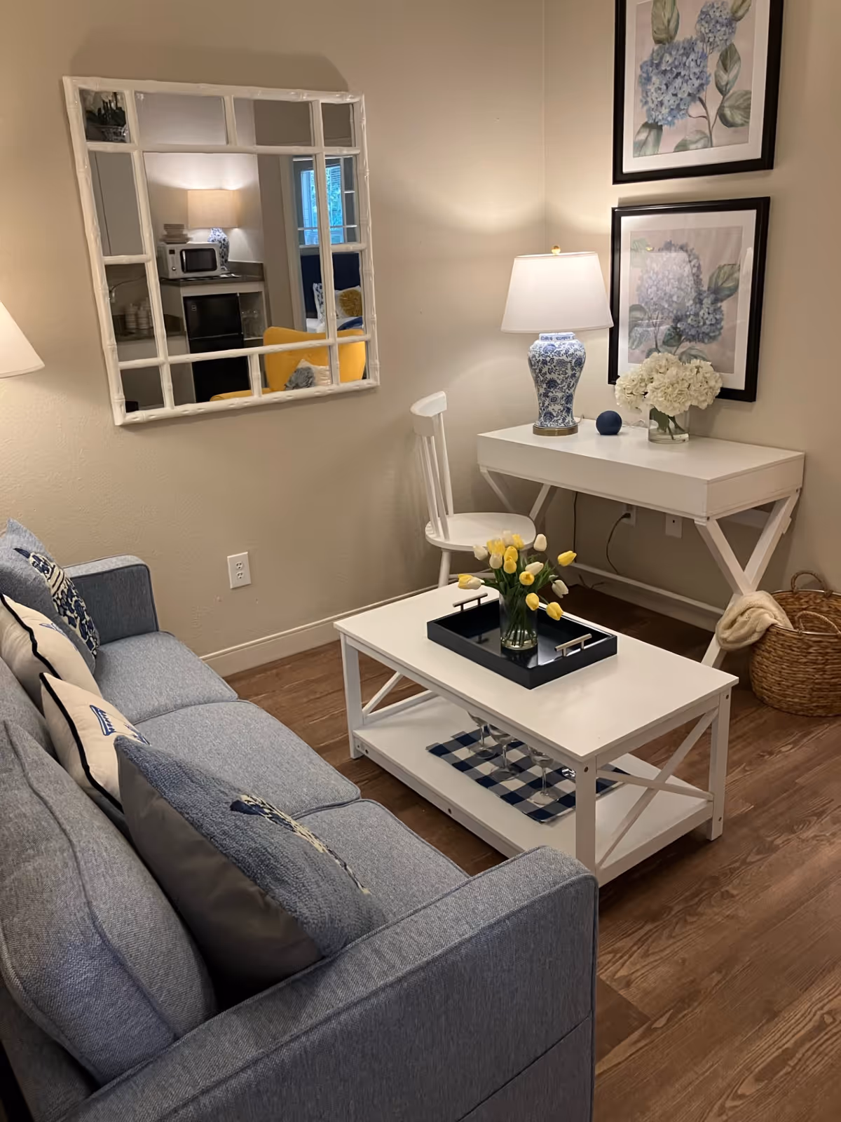 A cozy living room area with a blue sofa adorned with decorative pillows, a white coffee table with a vase of yellow and white tulips, and a white desk with a chair. The desk has a blue and white lamp, a small blue decorative ball, and a vase of white flowers. Two framed floral artworks hang on the wall above the desk. A large mirror on the adjacent wall reflects part of the room, including a yellow chair and a microwave on a counter. The floor is wooden, and a woven basket with a blanket is placed near the desk.