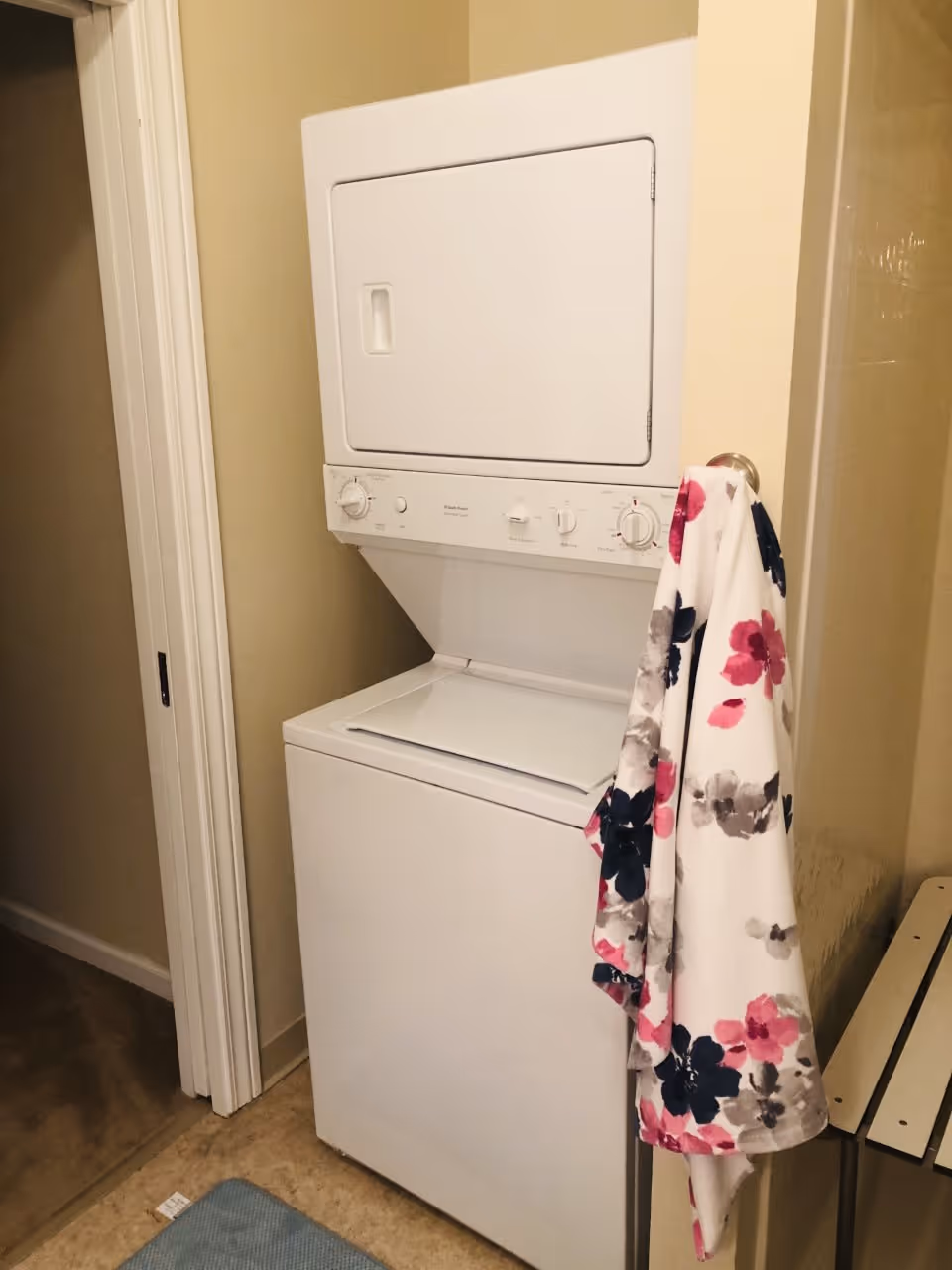 A stacked white washer and dryer unit in a small laundry area with beige walls. A floral-patterned towel with pink, navy, and gray flowers hangs on a hook attached to the wall next to the unit. A blue mat is on the floor in front of the washer and dryer.