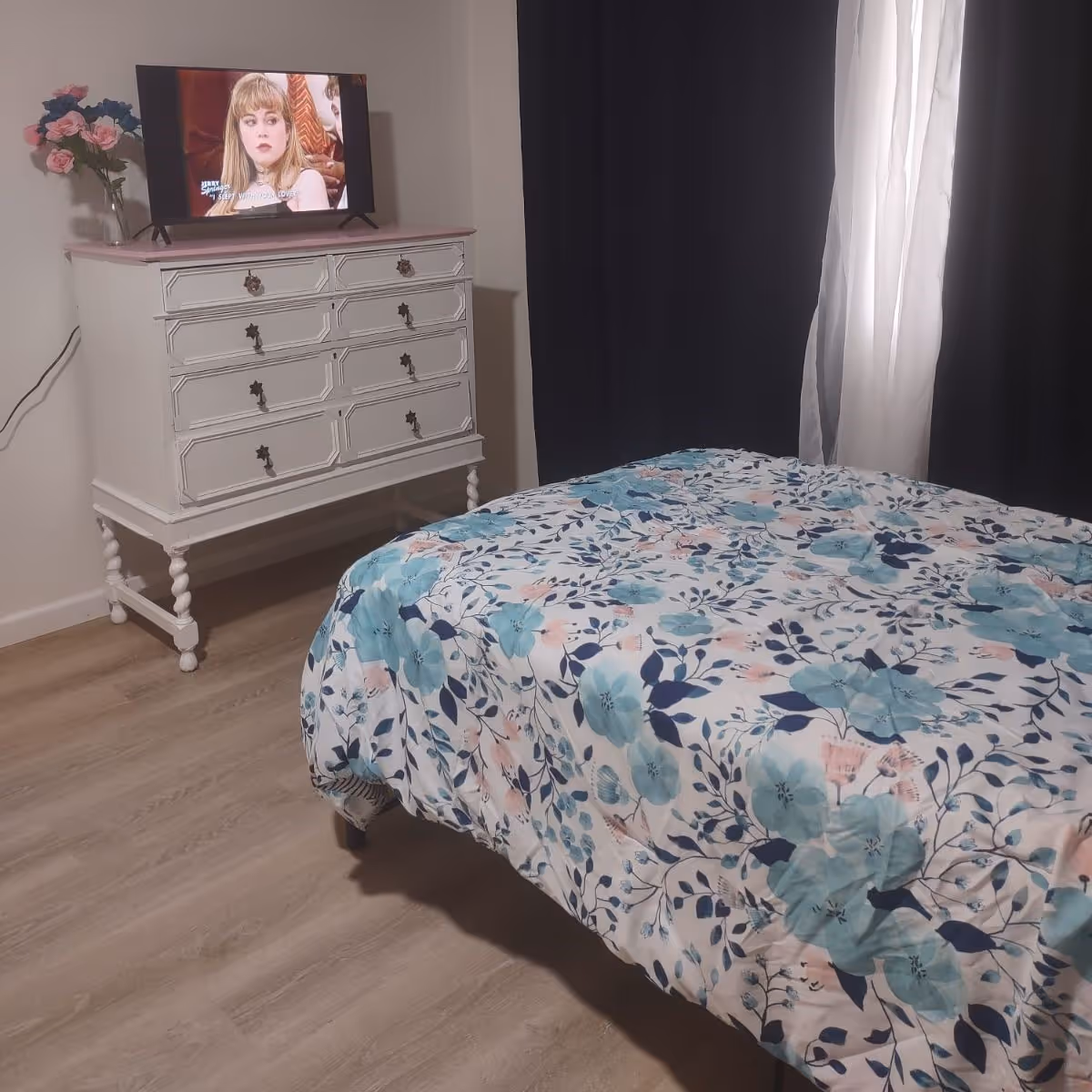 A bedroom with a floral bedspread, a white dresser holding a TV and vase of flowers, and dark curtains over a window.