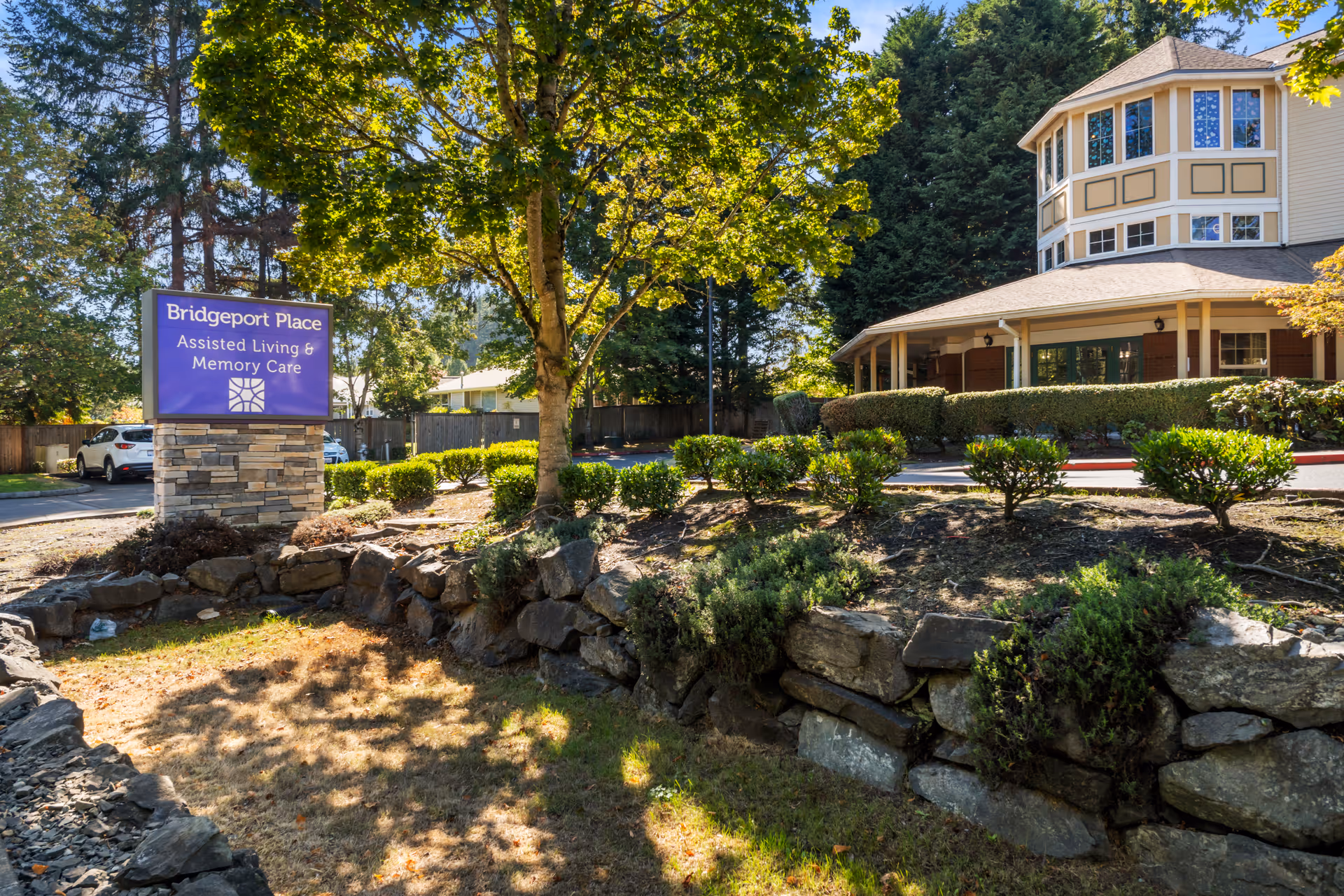 Exterior view of Bridgeport Place Assisted Living & Memory Care facility with a stone sign in the foreground surrounded by greenery and trees, and the building with large windows and a covered porch in the background.