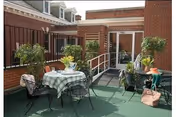 Sunlit outdoor patio with metal tables and chairs, potted plants, and a ramp leading to a doorway in a brick building.