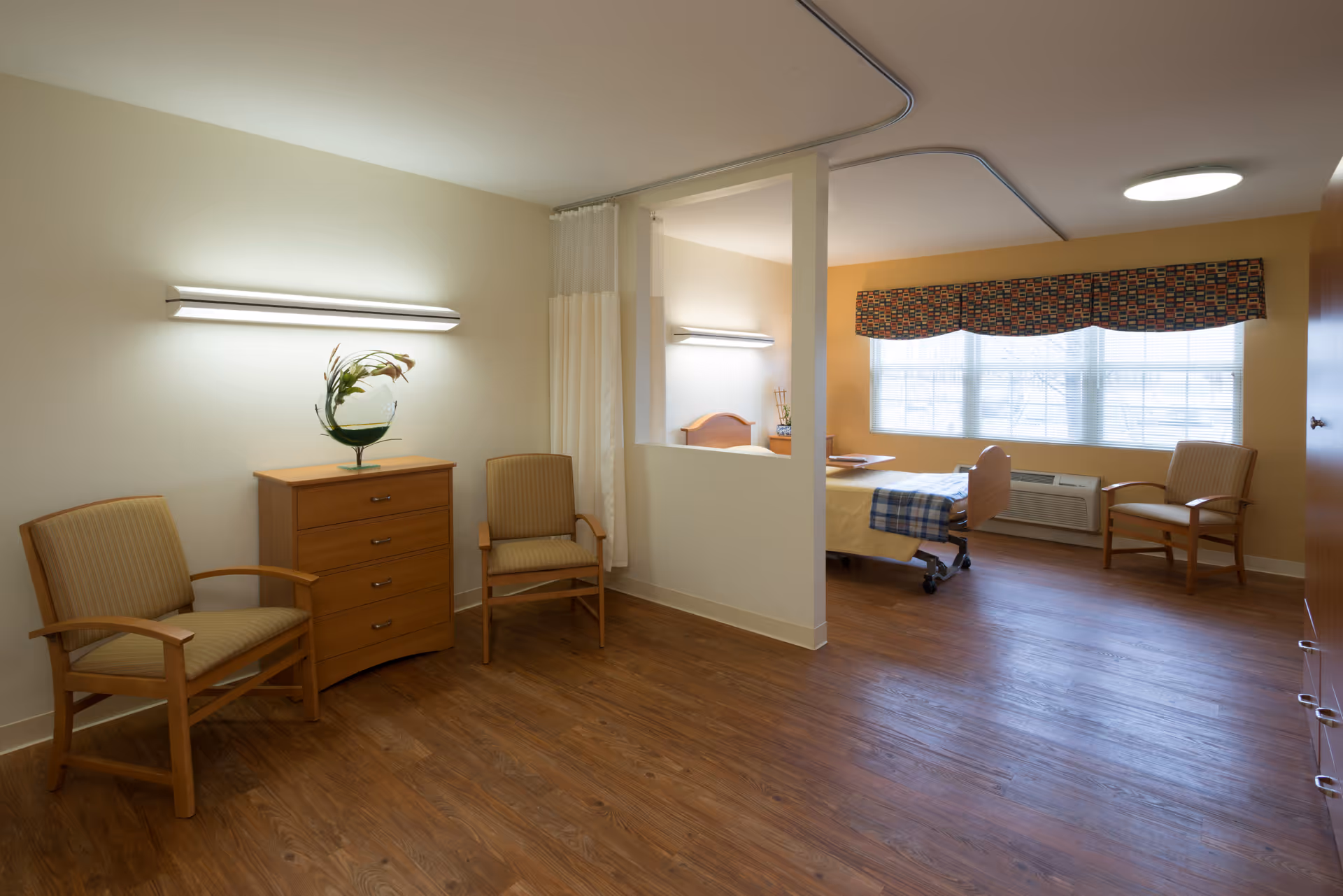 A spacious senior living bedroom with wooden flooring, two armchairs, a wooden dresser with a decorative plant, and a hospital-style bed near a large window with patterned valance. The room is divided by a half wall with a curtain for privacy.