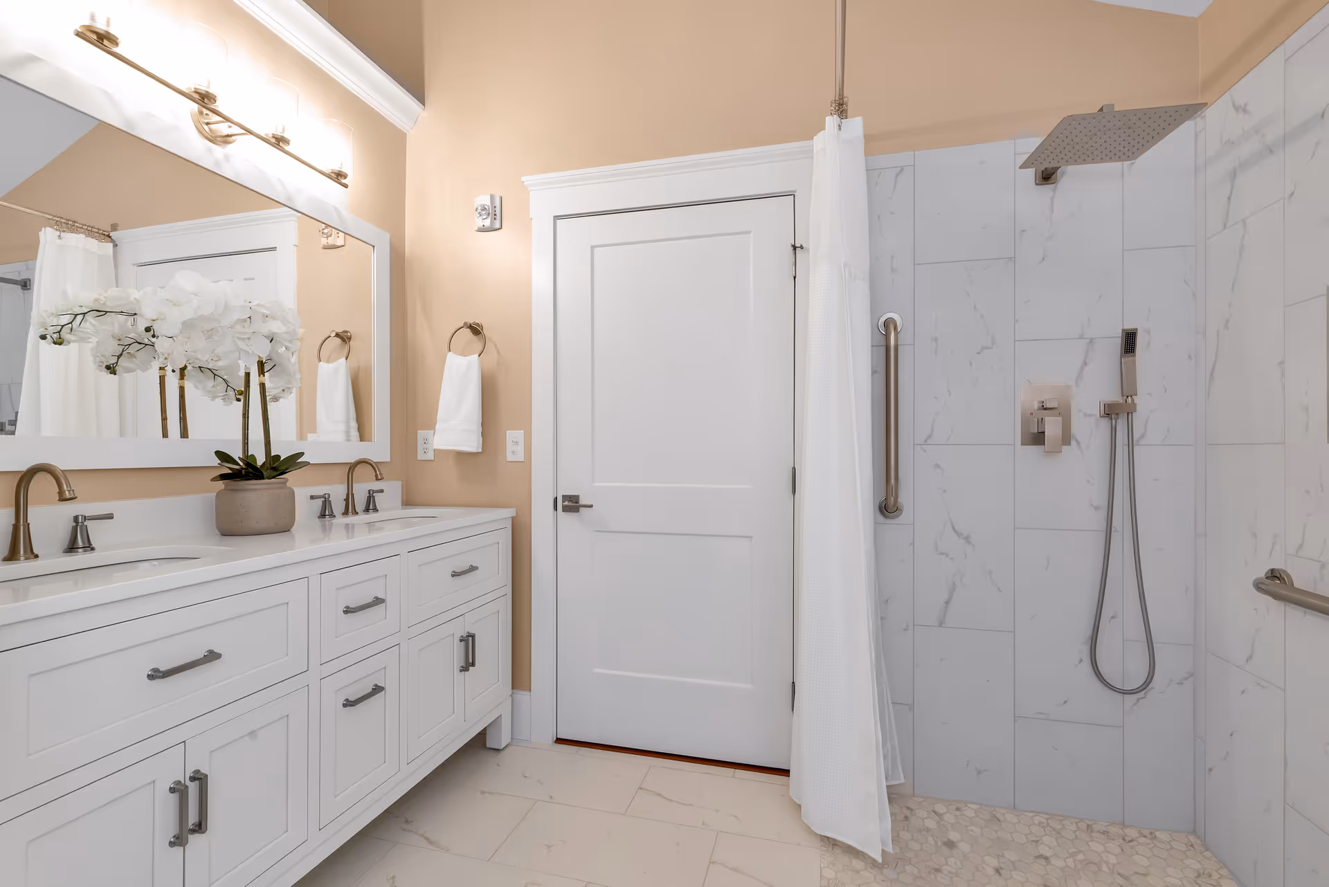 A modern bathroom with a double sink vanity featuring white cabinets and silver handles. Above the vanity is a large mirror with a light fixture. A potted white orchid is placed on the countertop. To the right, there is a walk-in shower with marble-like tiles, a rainfall showerhead, a handheld shower, and grab bars. A white shower curtain is partially drawn. The walls are painted beige, and the floor is tiled in a light color.