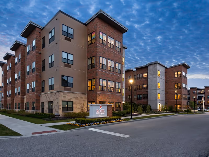 Dusk exterior view of a multi-story brick senior living building with lit windows and a sign reading 'Rose Senior Living Carmel'.