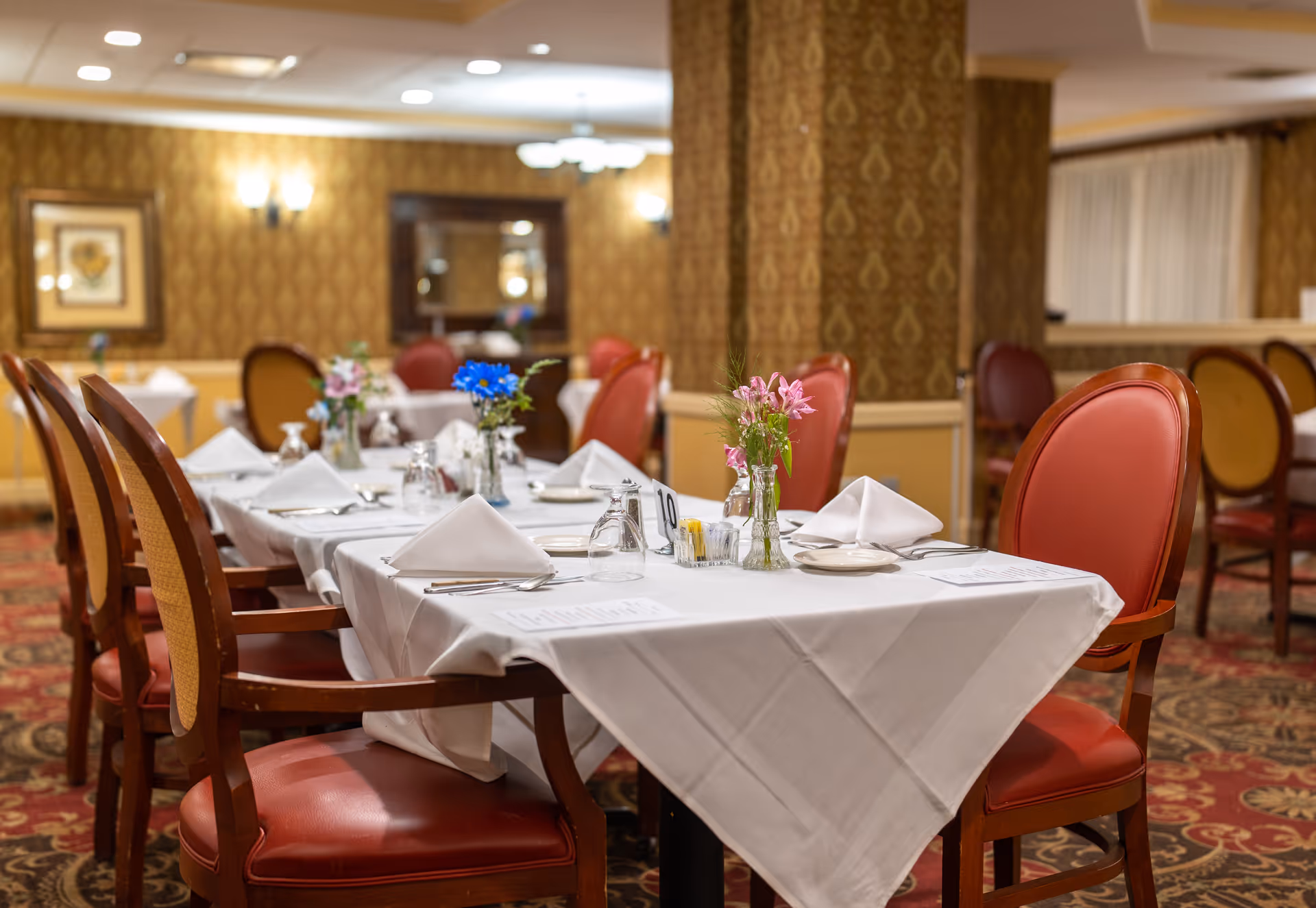 A dining room with tables covered in white tablecloths, set with folded napkins, plates, silverware, and small vases of flowers. The room has patterned wallpaper, carpeted floors, and wooden chairs with red cushioned seats and backs.
