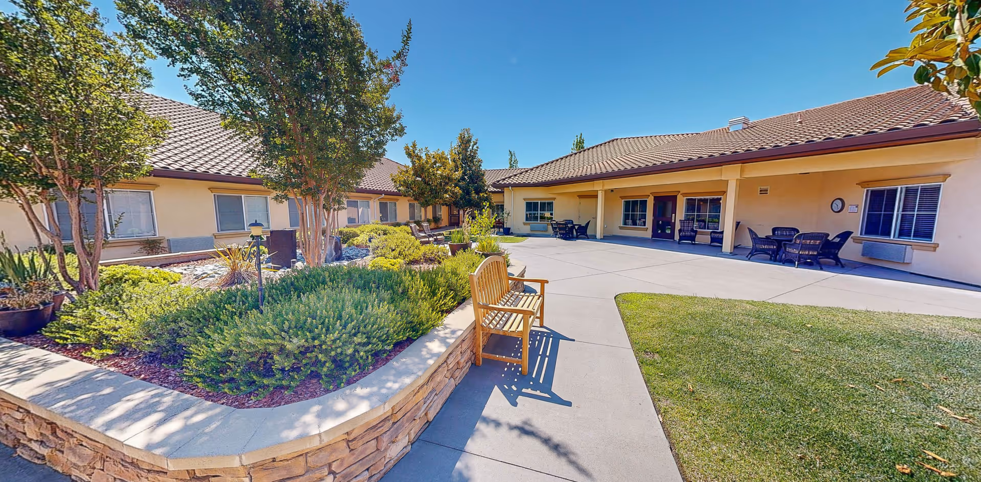 Outdoor courtyard area at Cogir of Folsom Memory Care featuring a stone planter with green shrubs and small trees, a wooden bench on a concrete pathway, and a building with a covered patio and outdoor seating under a clear blue sky.