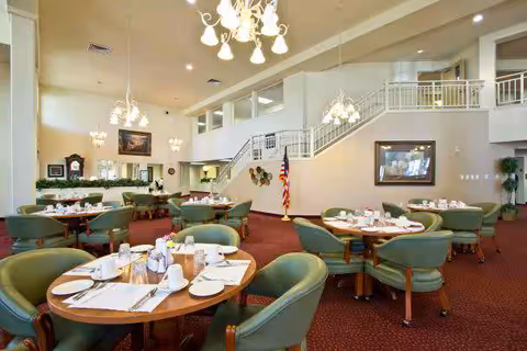 Spacious dining room with round tables set for service, green upholstered chairs, chandeliers, and an interior staircase.