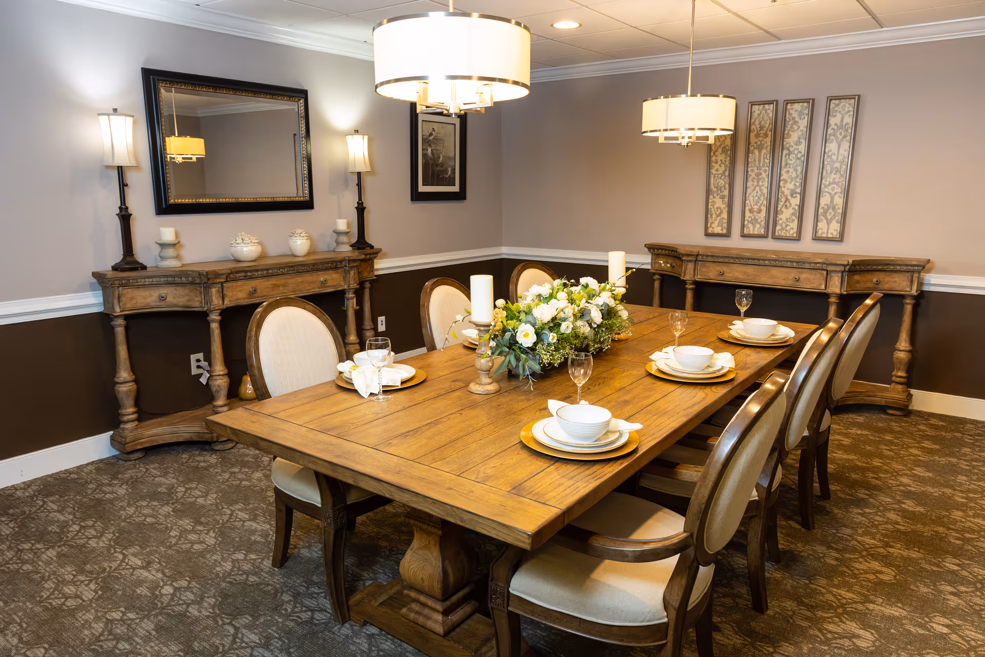 A formal dining room with a wooden table set for six, upholstered chairs, a floral centerpiece, pendant lights, and matching sideboards with decor.
