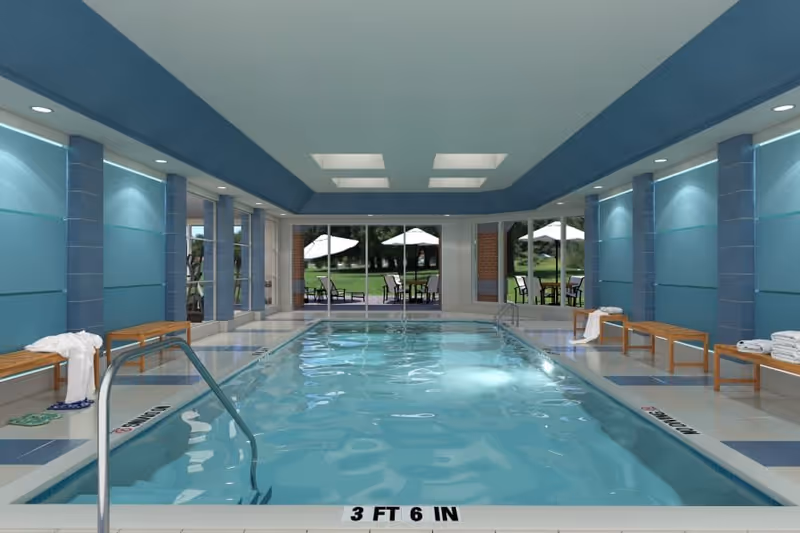 Indoor swimming pool with clear water, surrounded by blue walls and benches with towels. Large windows at the far end show an outdoor patio area with tables, chairs, and umbrellas, overlooking a green lawn and trees.