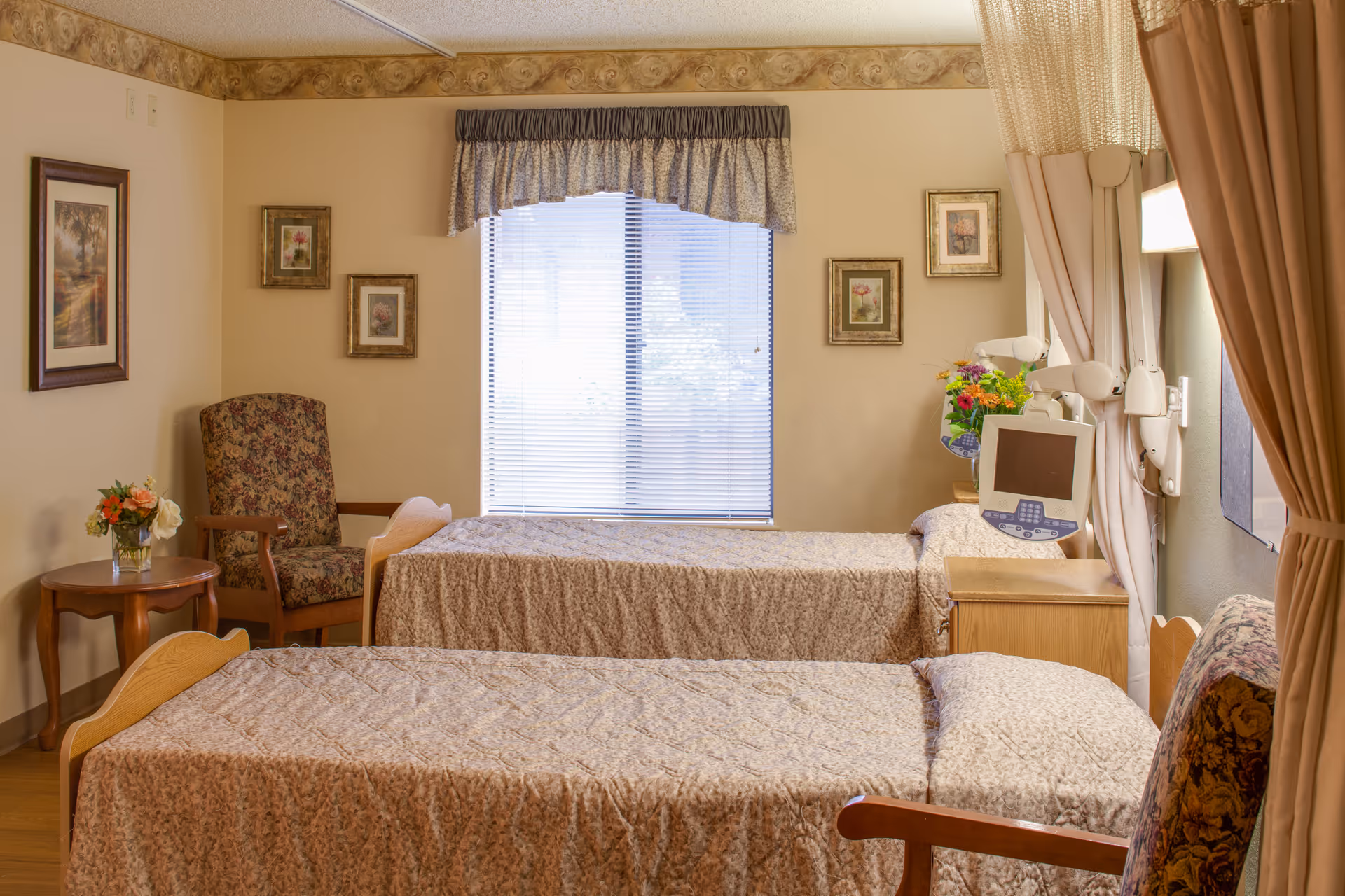 A cozy room with two single beds covered in beige patterned bedspreads. There is a floral armchair and a small wooden table with a vase of flowers. The room has a window with blinds and a valance, framed pictures on the walls, and medical equipment mounted near one bed. Beige curtains are partially drawn on the right side.