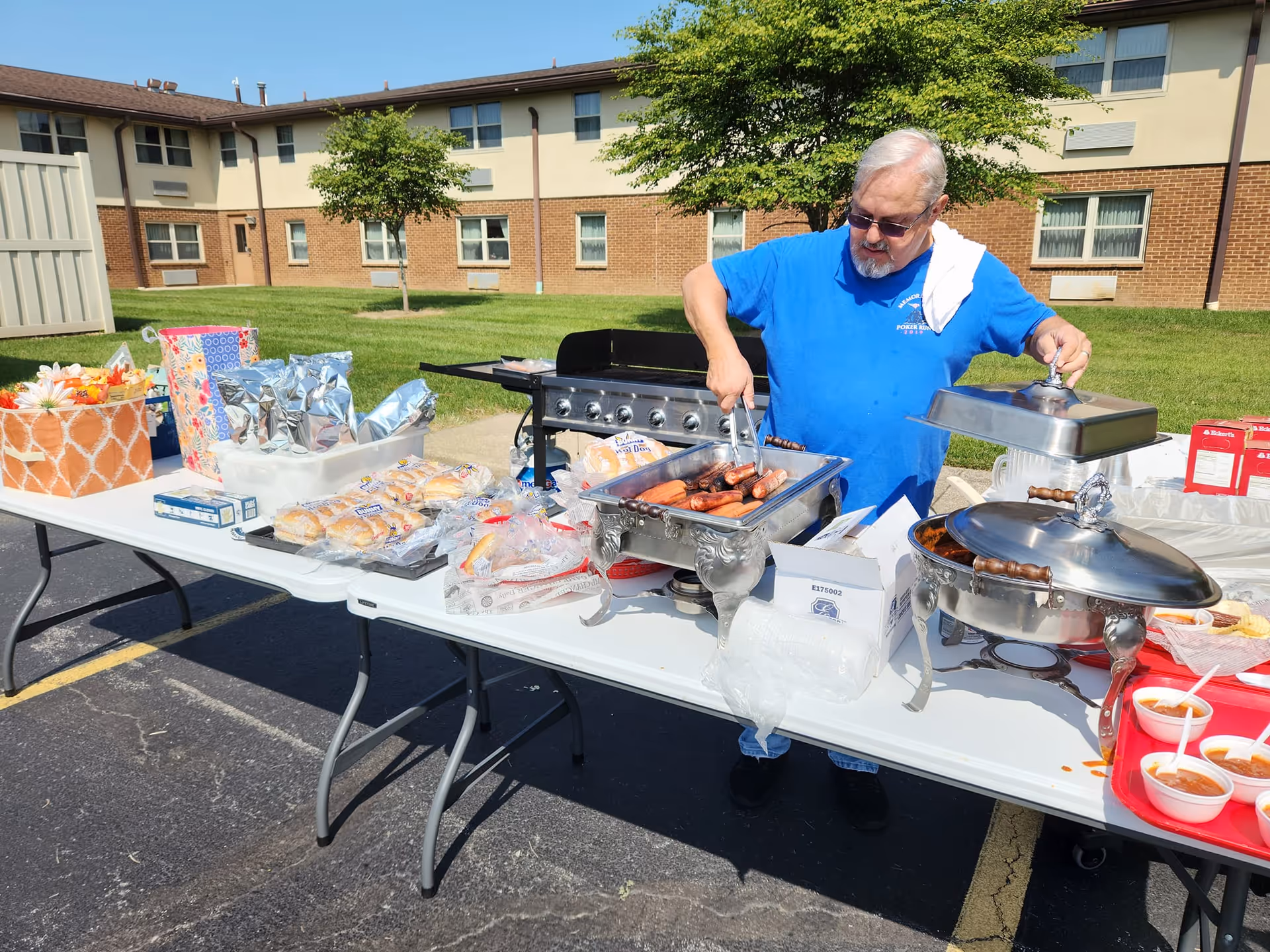 An older man wearing sunglasses and a blue shirt is serving grilled hot dogs from a chafing dish on a folding table outdoors. The table is set up with various food items including buns, wrapped packages, and cups of sauce. Behind the man is a large brick building with multiple windows and a grassy area with trees.