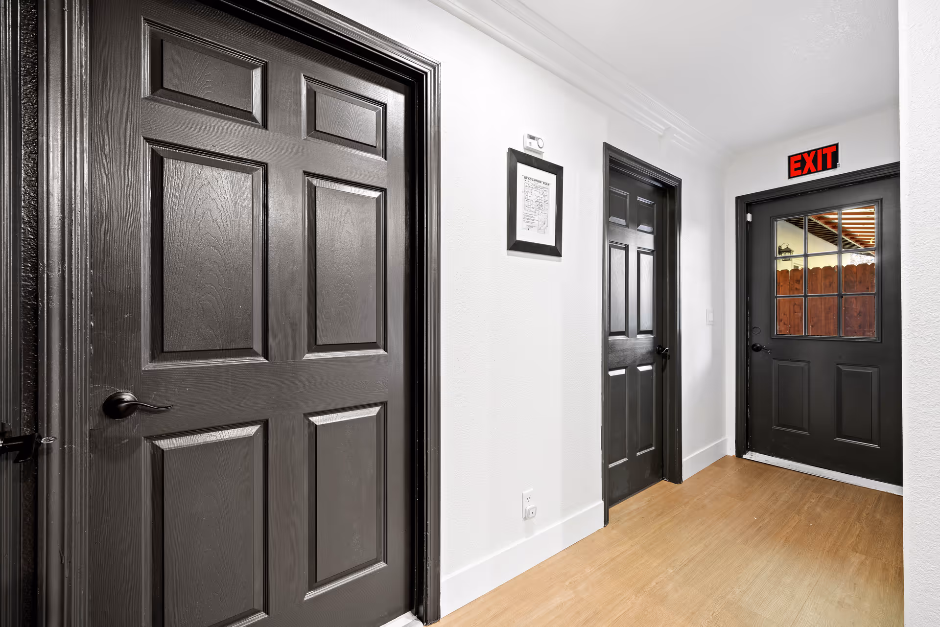 A hallway with three black doors and white walls. One door has a window with a view of a wooden fence outside and an illuminated red EXIT sign above it. The floor is light wood, and there is a framed document on the wall between two doors.