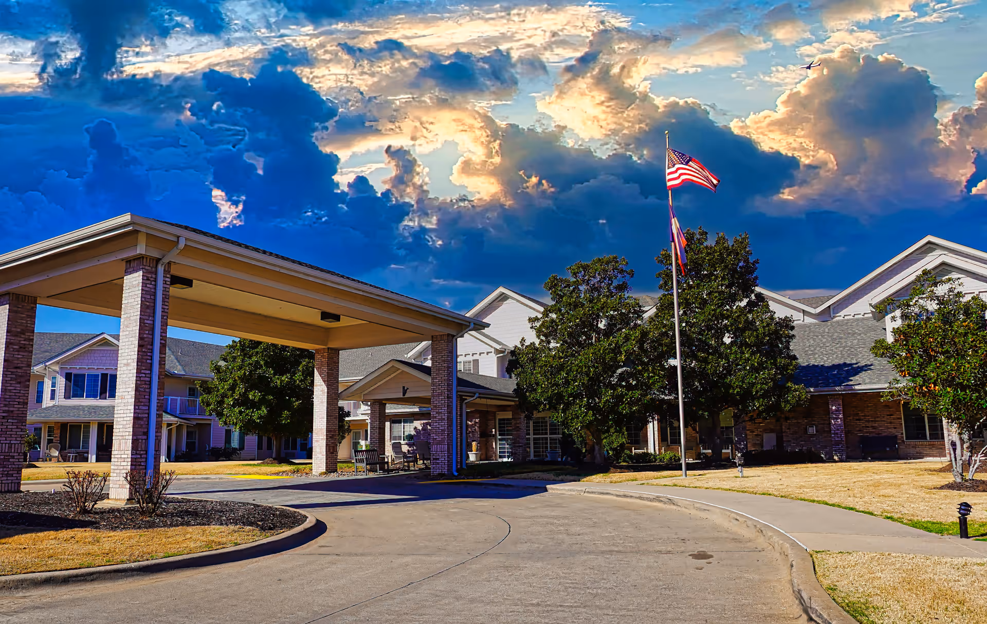 Exterior view of Pinewood Hills - Sky Active Living facility with a covered entrance, an American flag on a flagpole, trees, and a partly cloudy sky with dramatic lighting.