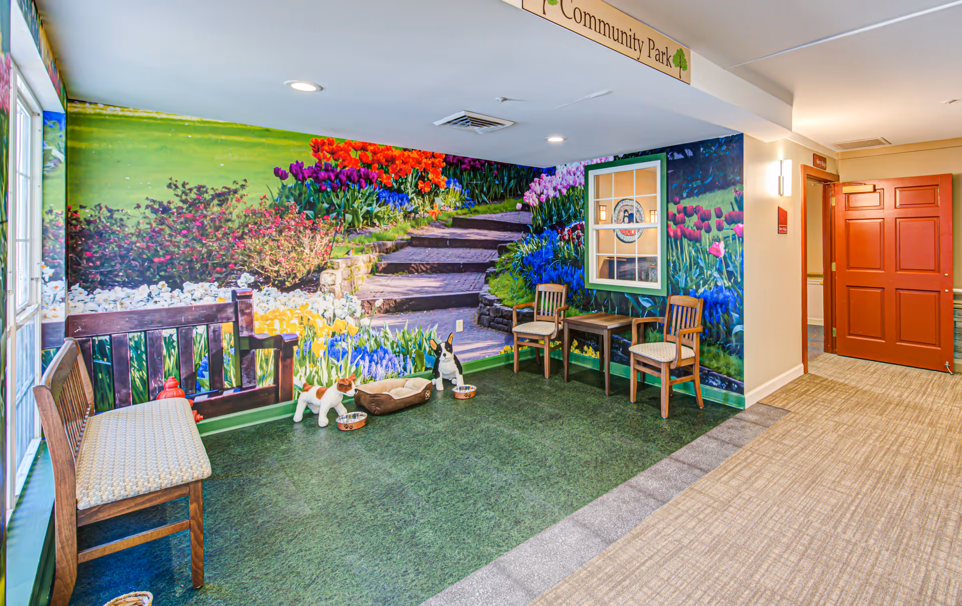 Indoor common area with benches and chairs facing a large colorful floral mural labeled 'Community Park,' with toy dogs and pet bowls on green carpet.
