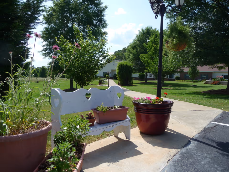 Outdoor garden area with a white wooden bench featuring heart-shaped cutouts, surrounded by potted plants and flowers. A paved walkway leads through a grassy area with trees and shrubs, and a building is visible in the background under a partly cloudy sky.
